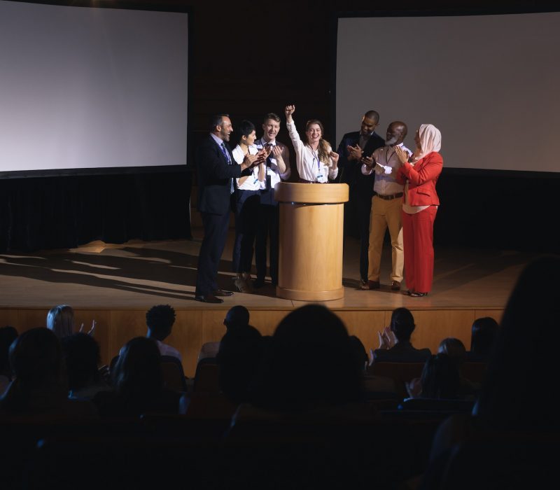 businesswoman-standing-at-the-stage-of-the-auditorium-with-colleagues-in-front-of-audience.jpg